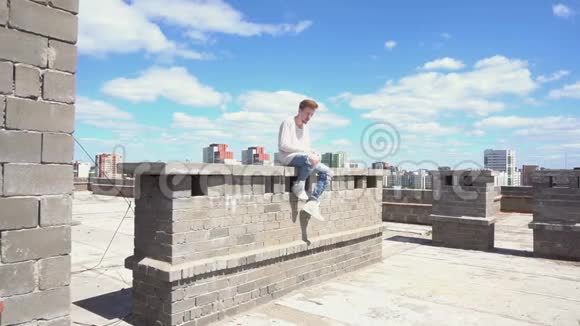 穿着白色运动衫的年轻红发男子坐在屋顶的砖墙上视频的预览图