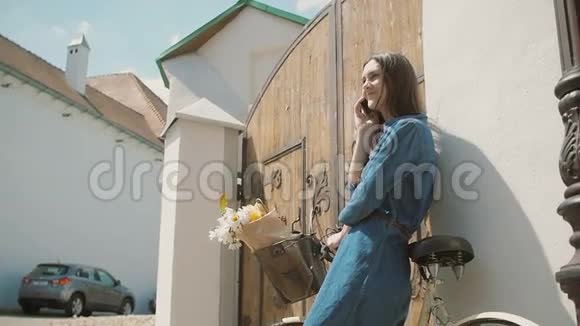 快乐的黑发女孩笑着打电话站在老门口花篮里放着自行车和鲜花慢吞吞的视频的预览图