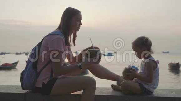 旅行者的母亲和女儿坐在海边在旅途中喝椰子视频的预览图