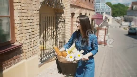 布鲁内特女孩微笑着对着镜头走着她的自行车带着鲜花和法国面包在篮子里慢节奏稳定的镜头视频的预览图