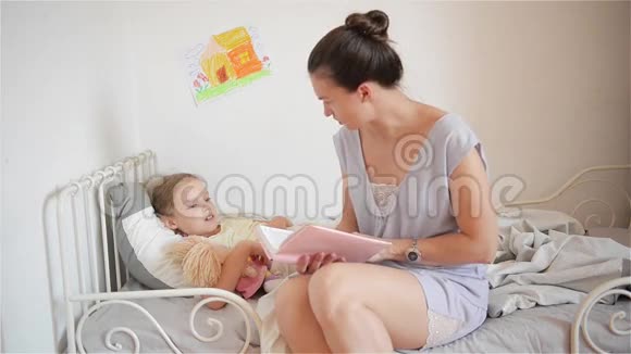 美丽的妈妈一边读童话一边让她的小女儿睡觉年轻的妈妈在床上读故事视频的预览图
