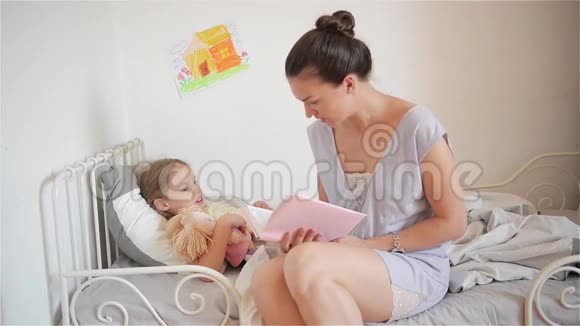 美丽的妈妈一边读童话一边让她的小女儿睡觉年轻的妈妈在床上读故事视频的预览图