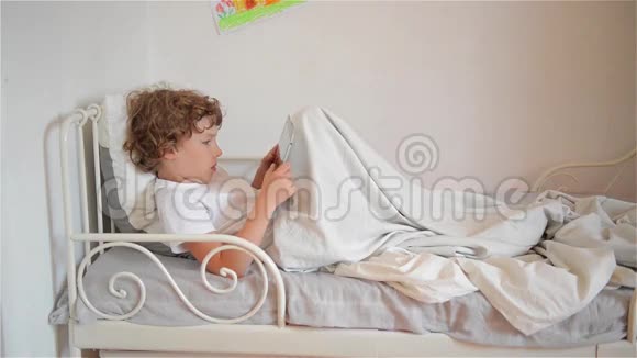 可爱的小男孩在床上玩数字平板电脑游戏视频的预览图