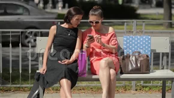 年轻漂亮的女孩坐在城市的公共汽车站两个女朋友在长凳上笑视频的预览图