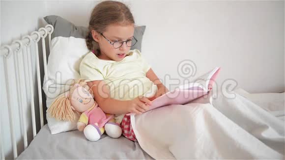 戴眼镜的小女孩躺在床上翻书也许孩子生病了她旁边坐着一个洋娃娃视频的预览图