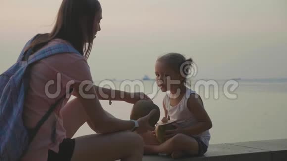 旅行者的母亲和女儿坐在海边在旅途中喝椰子视频的预览图