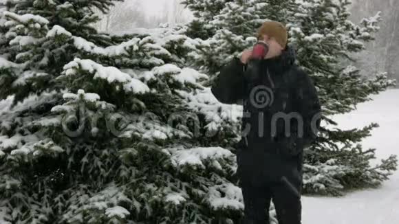 一个年轻人在冬天的森林里喝热水瓶里的热饮料大雪纷飞那个人在雪地里环顾四周视频的预览图