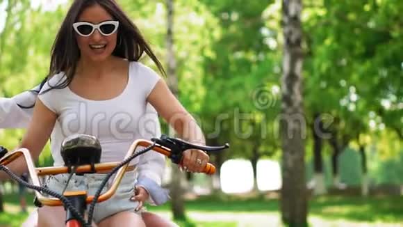 在公园里两个头发松松的年轻和黑发朋友穿着短牛仔短裤骑着电动摩托车骑着一辆电动摩托车视频的预览图