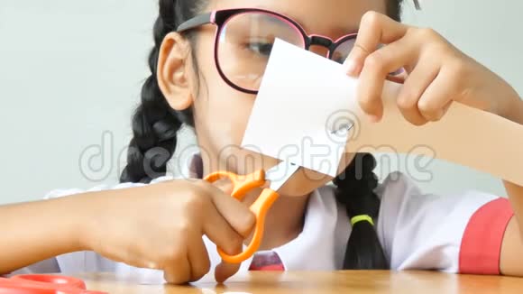 穿着泰国幼儿园校服的亚洲小女孩用剪刀在木桌上剪白纸视频的预览图