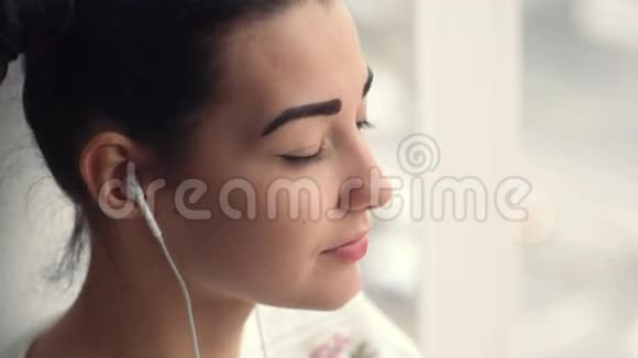 戴着耳机坐在窗边听音乐的迷人年轻女子视频的预览图
