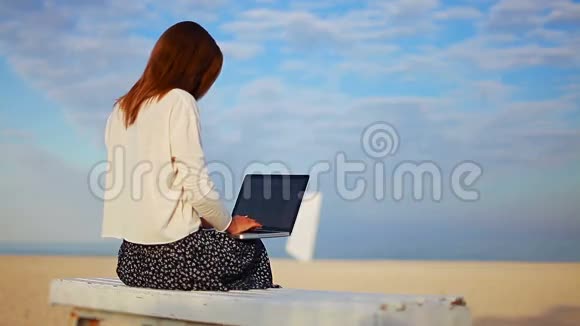 年轻女子坐在海边的长凳上使用笔记本电脑触摸板视频的预览图