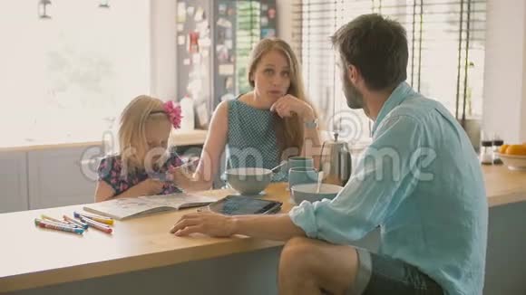 一位丈夫正在和他美丽的妻子说话而他们的女儿正在厨房的桌子上着色慢节奏视频的预览图