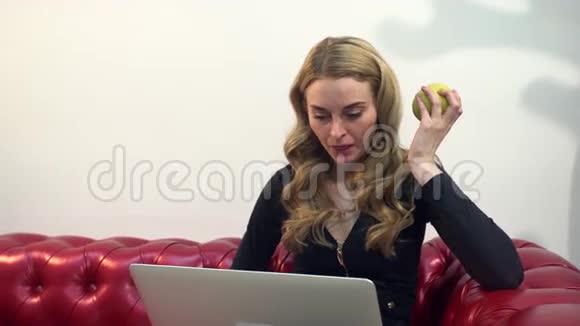 漂亮的金发美女在红色沙发上使用笔记本电脑并在客厅吃一个绿色苹果视频的预览图