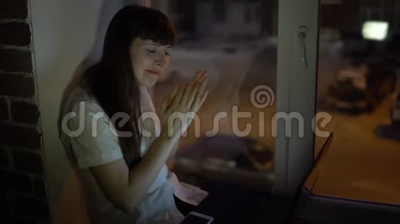 一个女商人坐在靠近一扇黑暗窗户的窗台上在笔记本电脑上工作视频的预览图