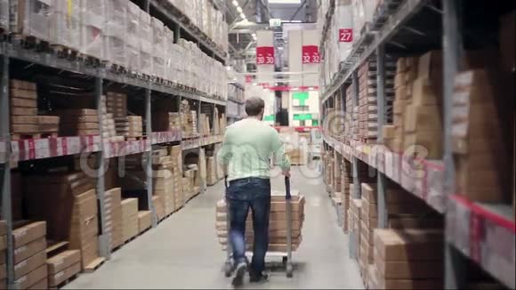 一个男人推着一辆装满箱子的手推车在仓库的货架之间视频的预览图