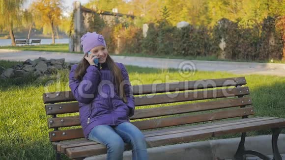 可爱的少女微笑着在秋公园坐在长凳上打电话牙套慢动作视频的预览图