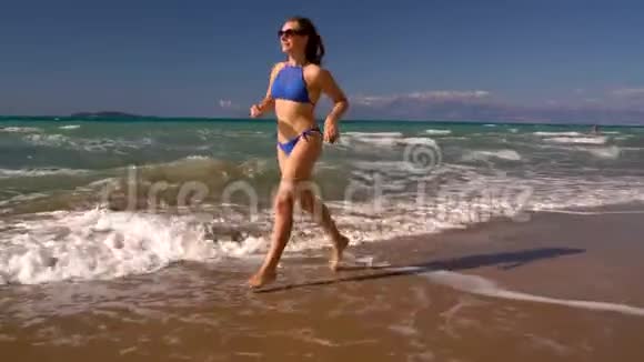 海滩比基尼女郎无忧无虑地沿着海滩上的水奔跑希腊科孚美丽的海岸视频的预览图
