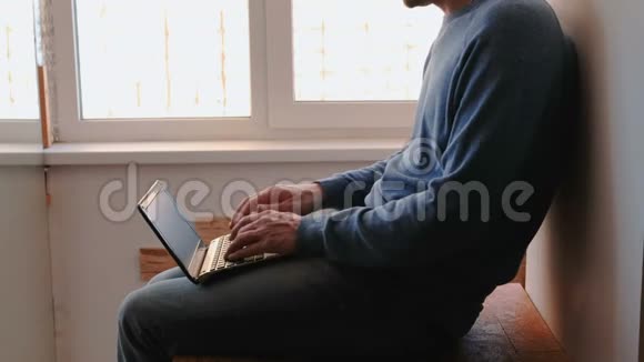 在笔记本电脑上工作一个面目全非的年轻人坐在靠近窗户的阳台上在笔记本电脑里打字视频的预览图