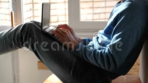 在笔记本电脑上工作一个面目全非的年轻人坐在靠近窗户的阳台上在笔记本电脑里打字视频的预览图