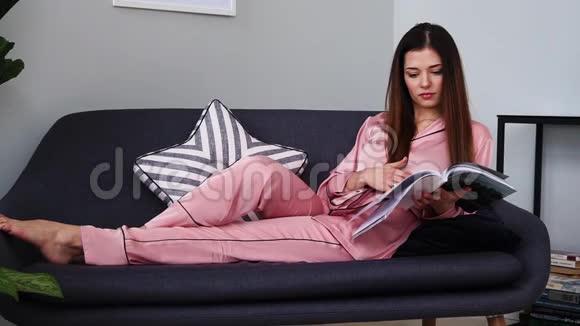 一个穿着粉红色睡衣的漂亮女孩看着一本光滑的杂志躺在沙发上从玻璃杯里喝香槟在视频的预览图