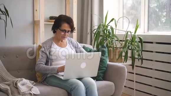 一位老年妇女正在家里使用坐在沙发上的笔记本电脑视频的预览图