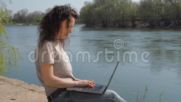 带着笔记本电脑在河边的年轻女子女孩坐在河岸上拿着笔记本电脑视频的预览图