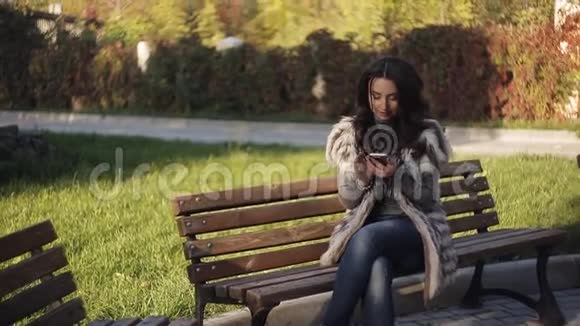 漂亮的女孩坐在你手机里的长凳上看照片凉爽的秋天公园视频的预览图