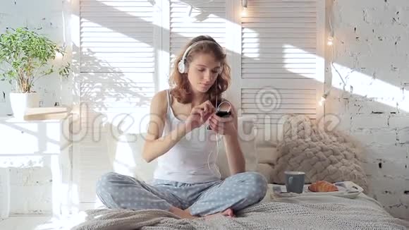 戴着耳机听着智能手机音乐在床上跳舞的快乐女人视频的预览图