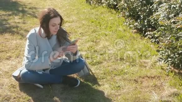 年轻漂亮的女人坐在草地上的春天公园里用手机说话视频的预览图