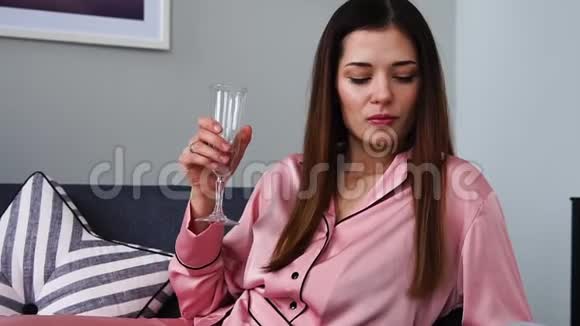 一个穿着粉红色睡衣的漂亮女孩看着一本光滑的杂志躺在沙发上从玻璃杯里喝香槟视频的预览图