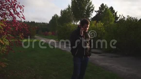 穿着黑色夹克的年轻人在秋天公园散步和打电话视频的预览图
