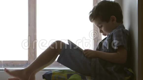 侧面图片可爱的小男孩在家的窗台上使用白色平板电脑视频的预览图