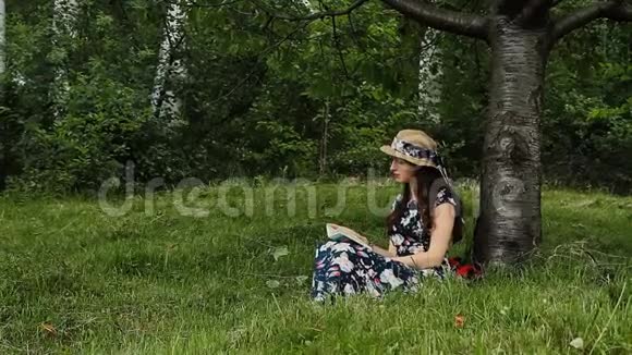 穿着衣服戴帽子的漂亮女孩正在农村度假时在树下看书视频的预览图