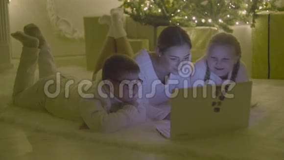 微笑的家人躺在圣诞树前的地板上看着笔记本电脑视频的预览图