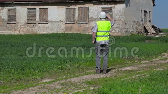 建筑督察员在草地上的旧废弃房屋附近拍摄平板电脑视频的预览图