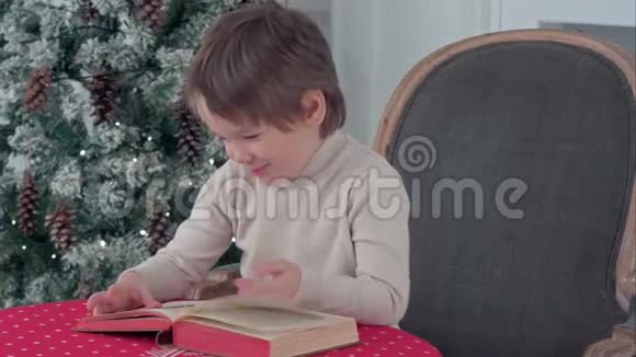 可爱的孩子看着书中的图画坐在圣诞树旁的椅子上视频的预览图