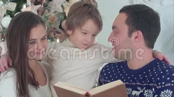 快乐的父亲读到圣诞老人的胡子而母亲抱着他们顽皮的儿子在圣诞树前视频的预览图