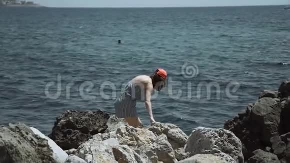 女孩站在海边的粗糙石头上看着海港那边的高楼大厦建设视频的预览图