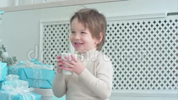 穿着白色毛衣的微笑男孩在圣诞树旁喝茶视频的预览图