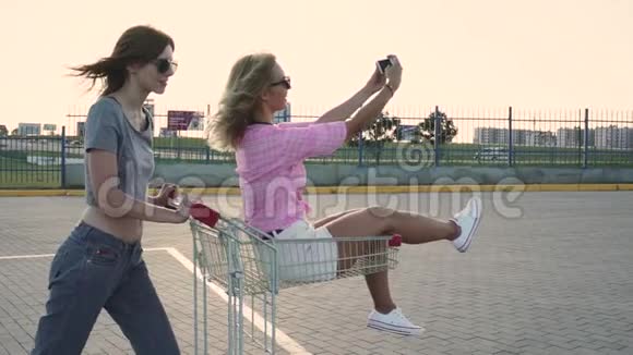 时尚有趣的年轻时髦女孩在购物中心停车场玩得很开心在街上的购物车里互相滚动慢动作视频的预览图