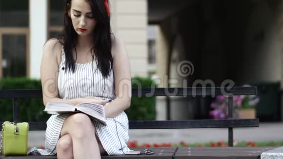 可爱的女孩头上有一个红色的边缘坐在公园的长凳上读一本书视频的预览图