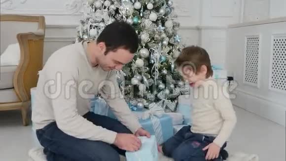 微笑的父亲和他快乐的儿子在圣诞树旁打开礼物视频的预览图