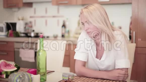 身着白衬衫的迷人的金发女郎坐在厨房的餐桌旁视频的预览图
