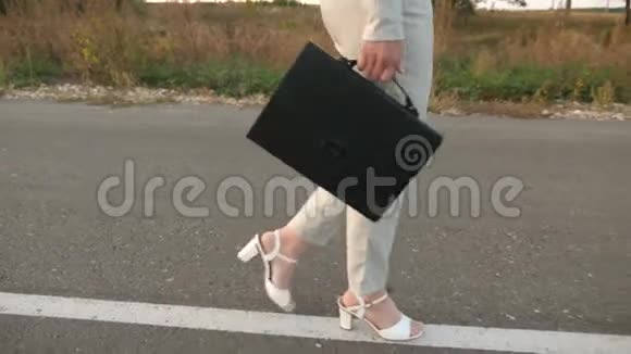 商务女性带着一个黑色公文包穿着轻便的西装穿着白色的高跟鞋沿着沥青路走视频的预览图