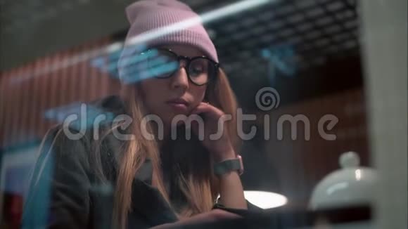 一位戴眼镜的时髦女人在咖啡馆里使用智能手机上的应用程序从窗口观看视频的预览图
