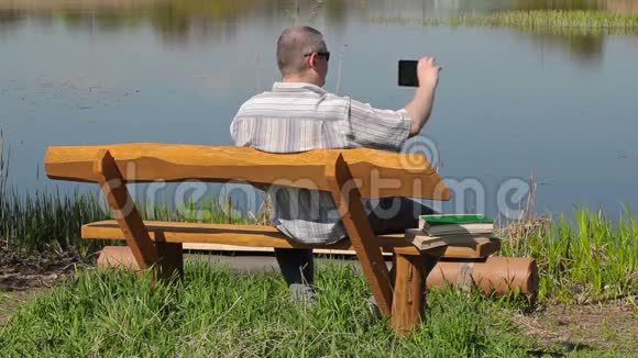 男人在河边的长凳上用智能手机拍照视频的预览图