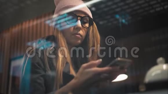 一个穿眼镜的时髦女孩在咖啡馆里使用智能手机上的应用程序从窗口观看视频的预览图