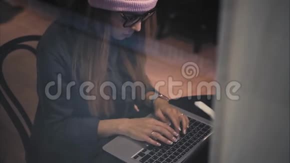一个时尚的时髦女性戴着眼镜和帽子在咖啡馆的笔记本电脑上打字从窗口俯视视频的预览图