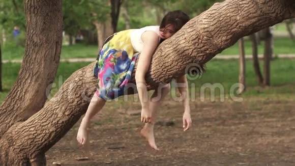 女孩很有趣躺在树枝上挂在她的手臂和腿上美丽的女孩在公园里做梦视频的预览图