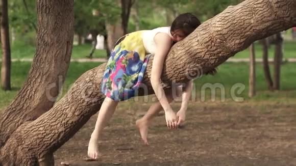 女孩很有趣躺在树枝上挂在她的手臂和腿上美丽的女孩在公园里做梦视频的预览图
